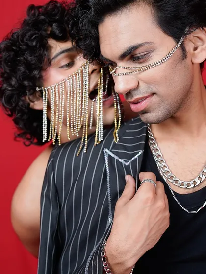 An intimate and edgy close-up of two male models with dramatic face jewelry. The red background and their intense connection create a powerful and high-fashion editorial photograph.
