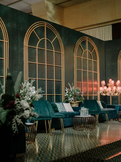 A luxurious lounge area from a "Velvet & Moody Luxe" themed event. The deep teal velvet sofas are set against a custom backdrop with gold-arched window frames, creating a sophisticated and dramatic seating space.