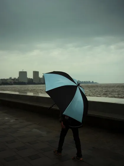 A person walks along the Marine Drive promenade, their two-toned umbrella adding a pop of color to a grey, overcast day. The image has a solitary and contemplative mood.