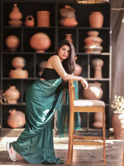 A modern take on saree styling. The model wears a teal saree paired with a contemporary black top, shot in a cafe with earthy tones. This shows how traditional wear can be styled for a modern, chic look.