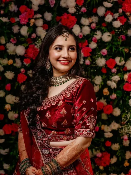 A happy bride against a floral backdrop. Her red velvet lehenga is paired with soft glam makeup and loose curls for a romantic reception look.