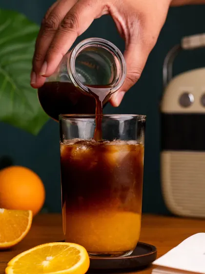 An action shot of espresso being poured over orange juice to create a unique coffee drink. The swirl of the dark coffee into the bright juice is visually captivating.