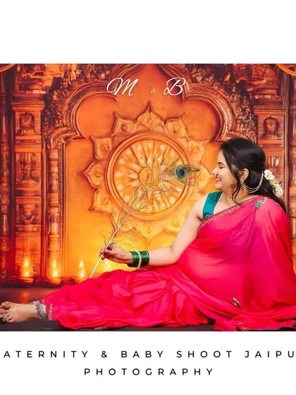 Celebrating tradition with a beautiful maternity portrait of a woman in a vibrant pink saree. She is seated gracefully against a warm, ornately decorated backdrop with diyas, holding a peacock feather in a classic cultural pose.
