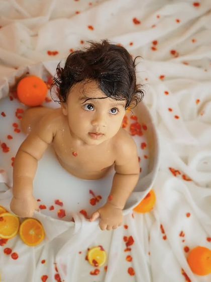 An overhead shot of a baby in a milk bath, looking up at the camera, surrounded by slices of oranges and red petals.