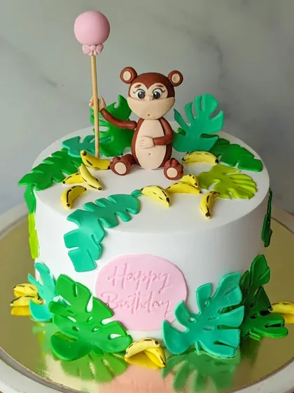 A cute monkey-themed cake with a monkey holding a balloon, surrounded by bananas and tropical leaves.