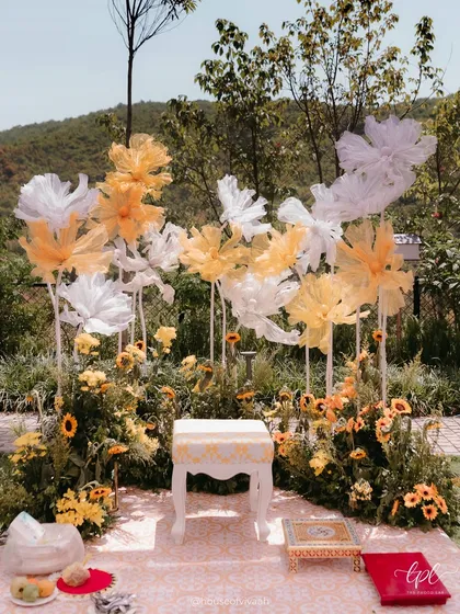The main seating for the Haldi ceremony, set against a backdrop of giant, ethereal paper flowers and a field of real sunflowers, creating a beautiful and artistic focal point.