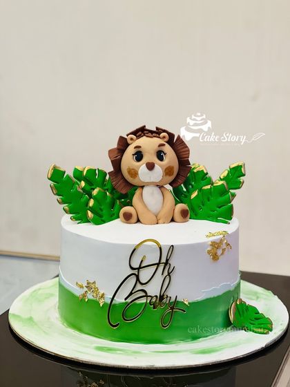 An adorable 'Oh Baby' shower cake with a jungle theme. This cake is topped with a cute fondant lion and decorated with green leaves and gold flakes.