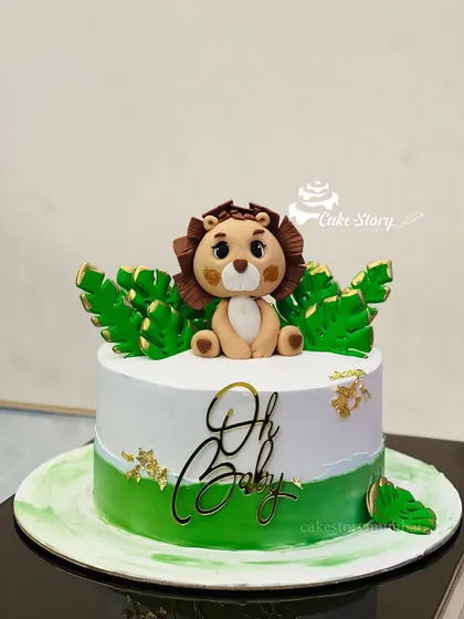 A cute jungle-themed 'Oh Baby' shower cake. This design features a sweet, handcrafted fondant lion cub sitting amongst tropical leaves.