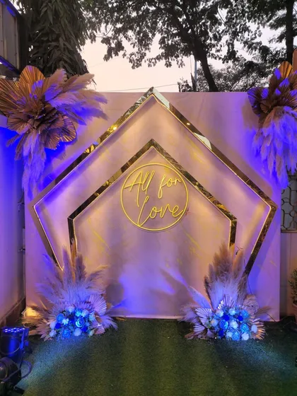 A modern bohemian photo booth with a blue-lit theme. The geometric gold frames, pampas grass, and "All for Love" neon sign create a stylish and contemporary look.