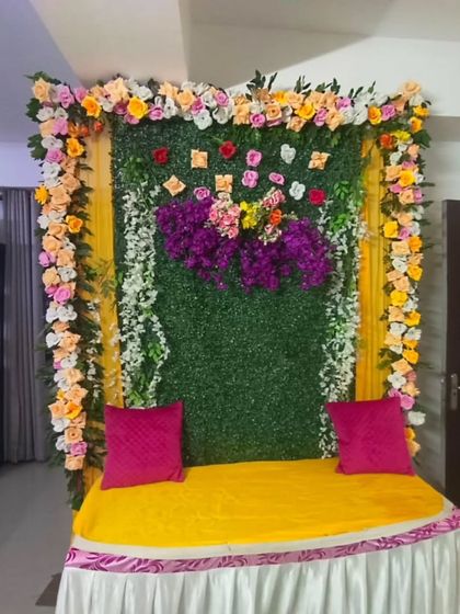 For intimate home functions, I create compact and beautiful backdrops. This one uses a green foliage panel, a frame of colorful flowers, and a comfortable gadda with bright pink cushions for the seating.