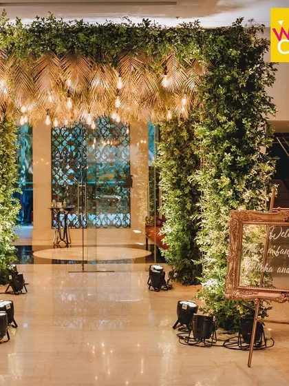Welcome to the party! For this Sangeet night, I created a dramatic tropical entrance with dense foliage and gold-painted palm leaves, lit by warm Edison bulbs to set an energetic and glamorous mood.