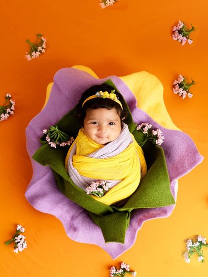 An adorable, smiling baby wrapped in yellow and lilac, nestled inside a prop that looks like a large, colorful flower.
