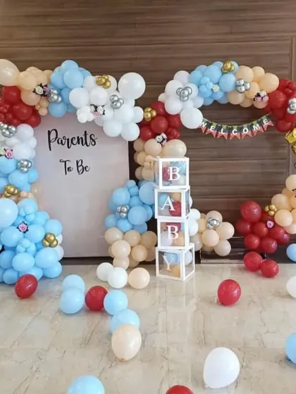 A "Parents To Be" baby shower decoration with a unique color scheme of red, blue, and nude balloons. The setup includes a custom backdrop, a balloon arch, and transparent "BABY" blocks.