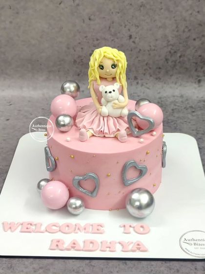 A cute doll cake to welcome a new baby. This pink cake features a sweet doll holding a teddy bear, surrounded by silver and pink decorative balls.