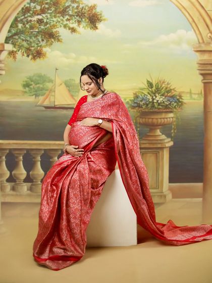 A stunning portrait of a mother-to-be in a rich red Banarasi saree. The painted backdrop and her graceful pose create a timeless and artistic image.