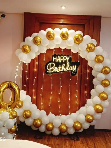 An elegant 50th birthday decoration for a home setting. A circular balloon arch in white and gold frames a doorway with fairy lights, complemented by large gold foil number balloons.