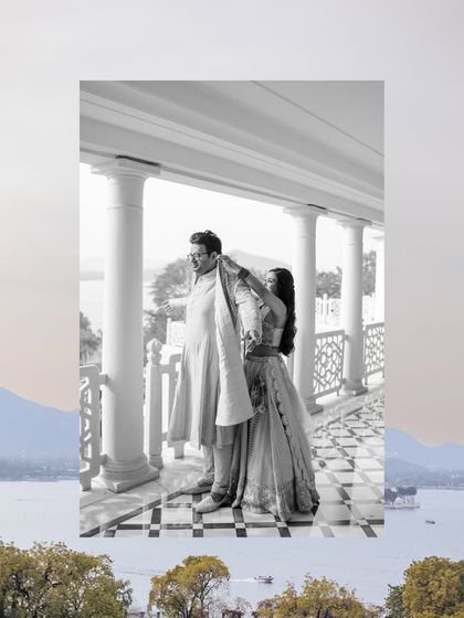 A creative black and white shot of the bride helping the groom with his outfit, framed by the stunning Udaipur landscape.