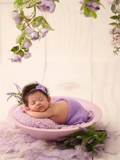 A baby in a purple wrap and headband sleeps in a bowl, with delicate lavender flowers hanging from above, creating a serene and color-themed scene.