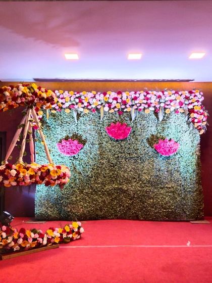 A classic naming ceremony setup with a crescent moon cradle decorated in red, yellow, and white flowers. The green wall backdrop is adorned with pink lotus accents.