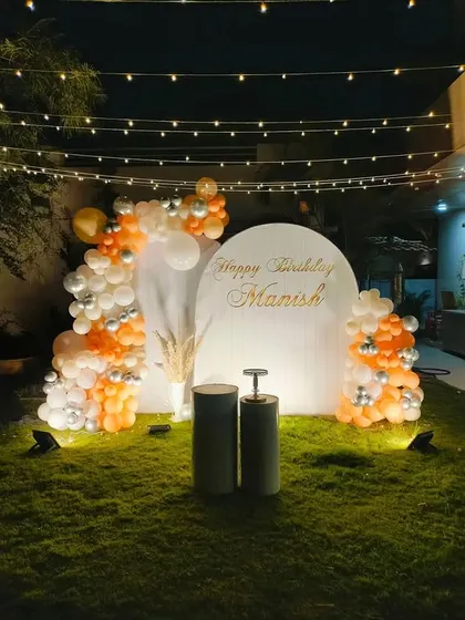 An elegant and simple garden birthday decoration with a white and orange balloon arrangement, a personalized backdrop, and string lights overhead.