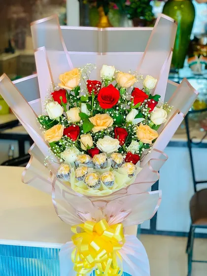 A beautiful hand-tied bouquet featuring a mix of red, white, and peach roses with Ferrero Rocher chocolates at the base. The arrangement is wrapped in sheer pink paper with a bright yellow bow.