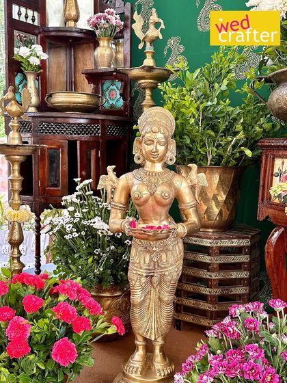 A brass 'Paavai Vilakku' (lady with a lamp) statue holding petals, surrounded by pink flowers and greenery, embodying South Indian tradition.