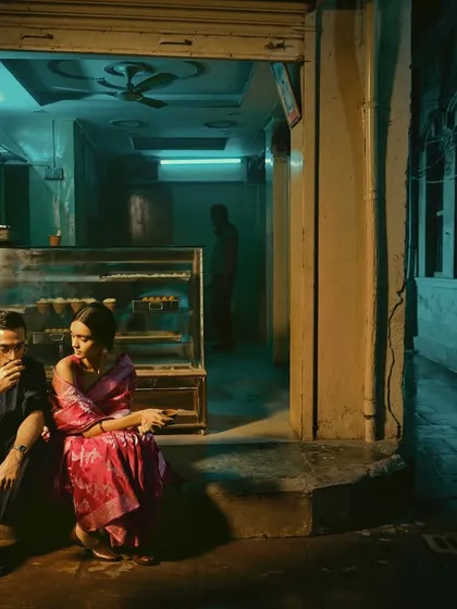 The couple sits outside a local sweet shop, bathed in the cool blue light of the evening. This shot grounds the cinematic story in a real, relatable setting.