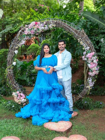 A happy couple posing in front of a large, rustic floral wreath in a garden. The mom-to-be's bright blue gown makes for a striking and joyful image.