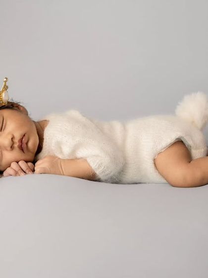 A sleeping newborn wearing a tiny gold crown and a white bunny outfit, posed on a plain grey background for a regal yet cute portrait.