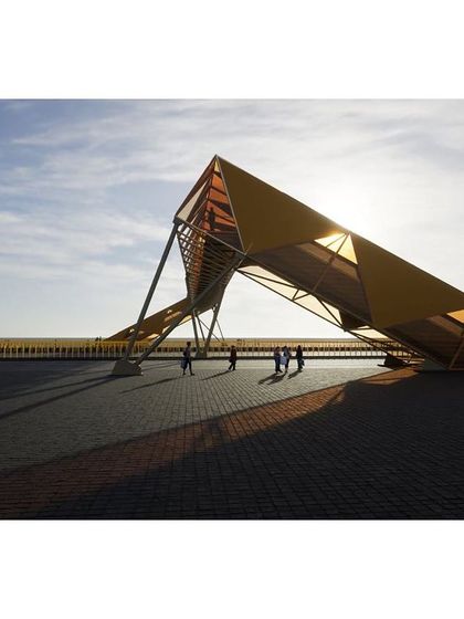 The dynamic, angular form of the footbridge creates a dramatic silhouette against the sky. The structure is designed to be experienced from multiple viewpoints, both by pedestrians and by drivers passing below.