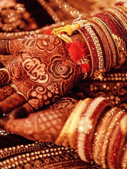 A close-up of a bride's hands, showing the deep maroon stain of the mehndi against her ornate dress and jewelry.