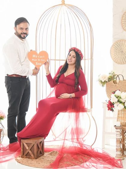 A happy couple's portrait in our bright and airy birdcage setup. The mom-to-be is seated in a beautiful red gown while her partner holds a sign celebrating their growing family.