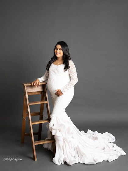 A beautiful portrait of a smiling mother-to-be in a white, long-sleeved lace gown with a ruffled train. She poses with a small wooden ladder, adding a rustic touch to the elegant shot.