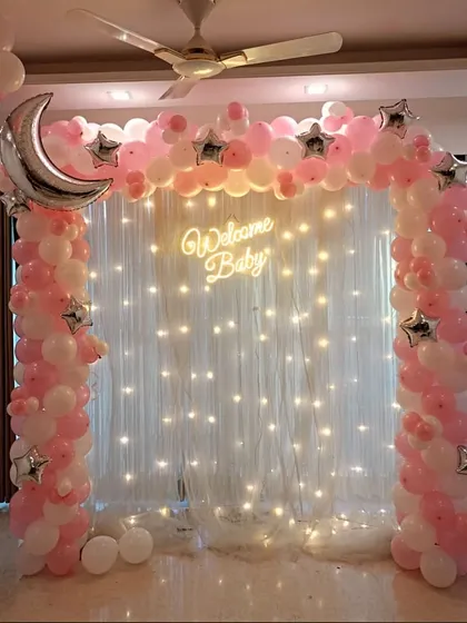 A dreamy 'Welcome Baby' setup perfect for bringing your little girl home. We created this magical moon and stars theme using a pink and white balloon arch, a large silver crescent moon foil balloon, and a backdrop of twinkling fairy lights for a truly special celebration.