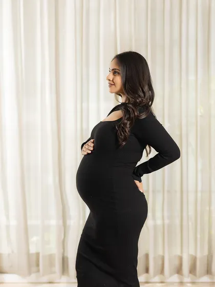 A classic and elegant maternity portrait. The mother-to-be, in a simple black dress, is beautifully silhouetted against a bright window, highlighting her form with grace.
