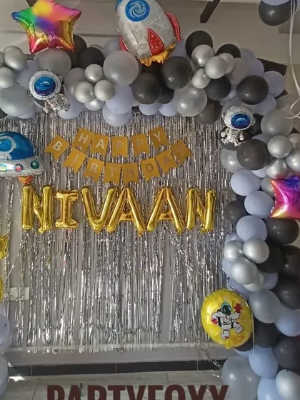 A space-themed 5th birthday decoration for a home party. This setup includes a silver fringe backdrop and a balloon arch with astronaut and rocket ship foil balloons.