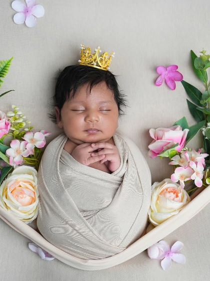 A newborn princess rests in a soft, cloth-lined hammock, crowned and surrounded by delicate pink and white flowers. This artistic setup creates a beautiful, fairytale-like portrait.