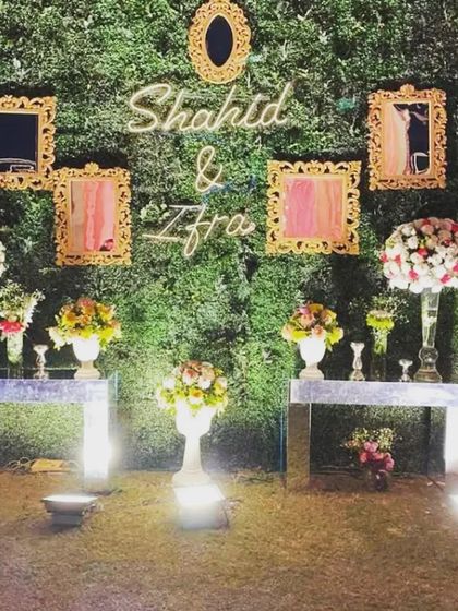 A creative selfie wall for a reception. The backdrop is a green wall decorated with ornate golden frames and a neon sign with the couple's names.