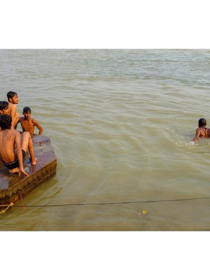 A group of boys sits on a stone platform by the Ganga, watching their friends swim. It's a simple, candid moment of friendship and leisure by the holy river.