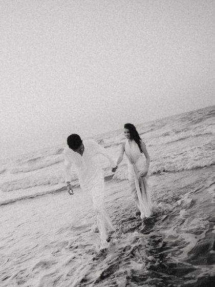 A candid black and white shot of the couple walking through the waves in Muscat. The image has a beautiful, grainy texture, capturing a spontaneous and joyful moment in a timeless style.