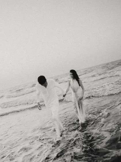 A candid black and white shot of the couple walking through the waves in Muscat. The image has a beautiful, grainy texture, capturing a spontaneous and joyful moment in a timeless style.
