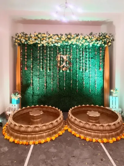 This Haldi setup features two large brass urlis for the ceremony, set against a green artificial grass wall with hanging floral strings and marigold borders.