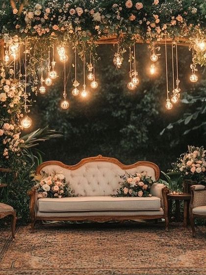 A rustic and romantic photo spot with a vintage sofa set under a canopy of flowers and Edison bulbs.