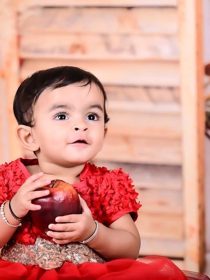 A close-up of a baby girl in a red dress holding an apple.
