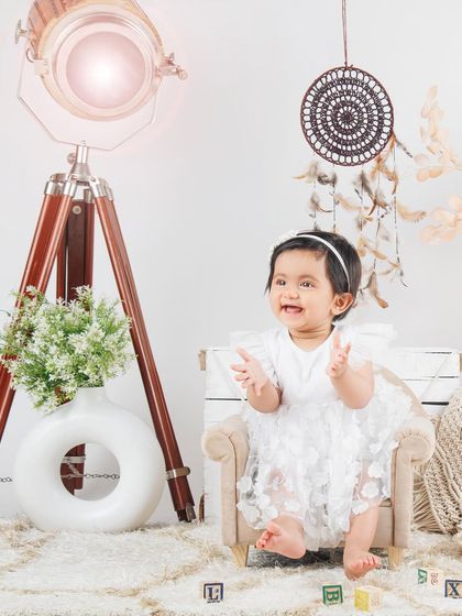 A classic portrait of a baby girl in a white dress, clapping her hands with joy.