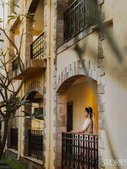 A bride stands on a balcony, gazing out as she awaits the start of her new journey. This portrait captures a quiet moment of reflection, her elegant figure framed by the building's architecture.