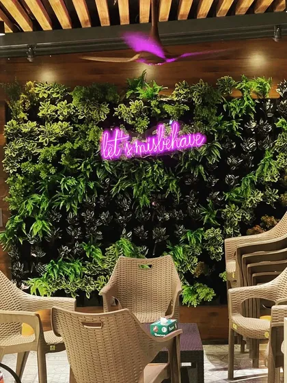 A living wall installation in a cafe, with a fun neon sign that reads "let's misbehave". The dark and light green plants create a lush, jungle-like backdrop for the seating area.