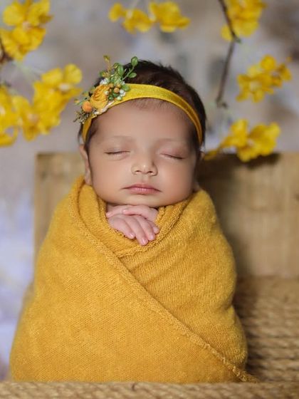 A newborn swaddled in a mustard yellow wrap sits propped up against a rustic wooden background with delicate yellow blossoms.