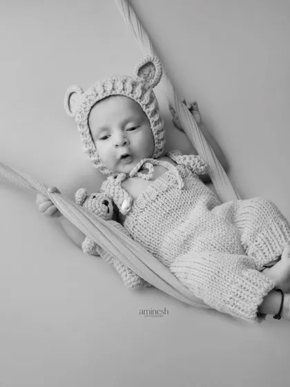 A timeless black and white portrait of a 20-day-old baby in a knitted bear outfit, holding a tiny teddy bear. The baby rests in a soft sling, creating a feeling of comfort and security.