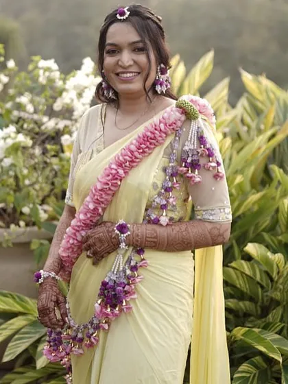 This bride chose a beautiful floral garland style for her Haldi. The long strand of pink and purple flowers worn over the shoulder adds a unique and graceful touch to her simple yellow saree.